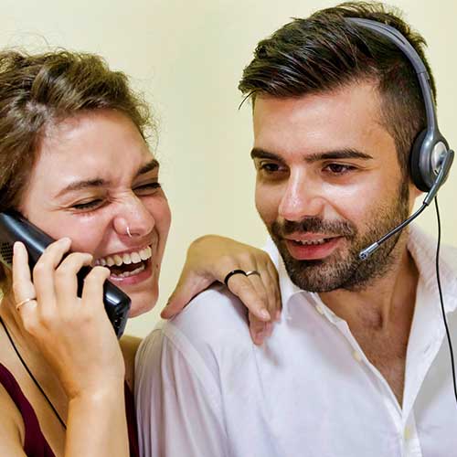 woman using a phone and man using a headset, both are laughing